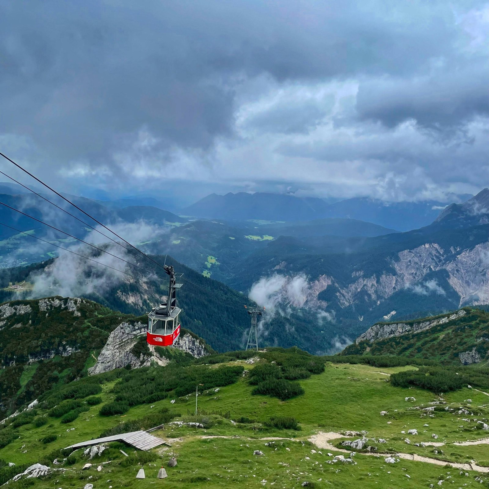 A red cable car traverses the lush Bavarian Alps under a cloudy sky.